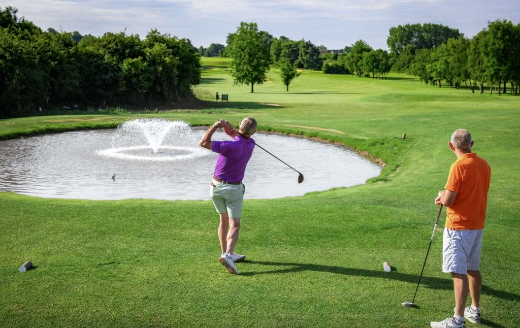 two men hitting over water fountain towards green at golf course in bristol