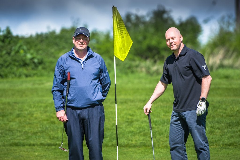 Two men smiling next to a golf flag on the green at a golf club in Bristol, perfect for golf days, golf lessons, and golf membership in Bristol.